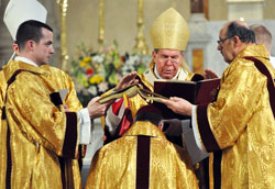 Archbishop Daniel M. Buechlein prays over Bishop-designate Christopher J. Coyne as transitional Deacon Dustin Boehm of Greenwood, left, and Deacon Joseph Holderried of St. Margaret Mary Parish in Westwood, Mass., hold a Book of Gospels over the new auxiliary bishop&rsquo;s head on March 2 at St.&nbsp;John the Evangelist Church in Indianapolis. This part of the ordination is a sign that the faithful preaching of the word of God is the pre-eminent obligation of the office of the bishop. (Photo by Mary Ann Wyand)
