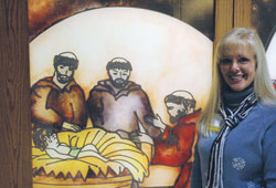 For 17 years, Shirley Fox has planned the Live Nativity at Franciscan St. Francis Health&mdash;Beech Grove. This year, the annual event will be held on Dec. 12. Here, Fox poses by a glass Nativity scene of the Baby Jesus being admired by Franciscan friars. The artwork is outside the chapel of Franciscan St. Francis Health&mdash;Indianapolis. (Photo by John Shaughnessy)