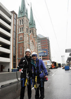 Benedictine Sisters Jennifer Mechtild Horner, left, and Sheila Fitzpatrick pose under the 800-foot zip line they rode on Jan. 26 that was built over Capitol Avenue in Indianapolis in the middle of the&nbsp;Super Bowl Village next to St. John the Evangelist Church. Both sisters are members of Our Lady of Grace Monastery in Beech Grove, which is hosting the workers that constructed and are operating the zip line. (Photo by Sean Gallagher)