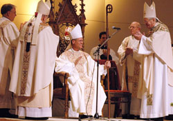 Bishop Paul D. Etienne, center, sits in the bishop&rsquo;s chair, also known as the cathedra, of the Diocese of Cheyenne, Wyo., on Dec. 9, 2009, at the Cheyenne Civic Center during his ordination and installation as the shepherd of the Church in Wyoming. The principal action of installing a bishop as the spiritual leader of a diocese is the seating of him in the cathedra. Applauding Bishop Etienne are, from left, Archbishop Charles J. Chaput, then of Denver, and Archbishop Pietro Sambi, then apostolic nuncio to the United States. Bishop Etienne was previously a priest of the Archdiocese of Indianapolis. (File photo by Sean Gallagher)