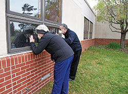Therese Brandon, left, and Father Joseph Moriarty visit their father, Patrick Moriarty, on April 30 through the window of their father&rsquo;s room at the St. Paul Hermitage retirement community in Beech Grove. For more than two months, visitors have not been allowed except for end-of-life situations to help protect the residents and staff members from the coronavirus. (Photo by Sean Gallagher)