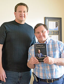 Greg, left, and Mark Hublar pose in Mark&rsquo;s New Albany apartment with the book Greg wrote about the against-the-odds accomplishments and impact of his brother, who overcame challenges of Down syndrome to become a motivational speaker advocating around the state and nation for the employment of those with disabilities. (Photo by Natalie Hoefer)