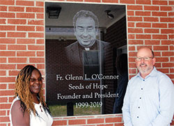 The smiling image of the late Father Glenn O&rsquo;Connor greets people near the entrance of the new Seeds of Hope residence center on the grounds of St. Joseph Parish in Indianapolis. The archdiocesan priest started the Seeds of Hope program in 1999 to help women overcome their struggles with drugs and alcohol. Standing near the tribute to Father O&rsquo;Connor are Marvetta Grimes, the executive director of Seeds of Hope, and Sean O&rsquo;Connor, one of the priest&rsquo;s seven siblings. (Photo by John Shaughnessy)