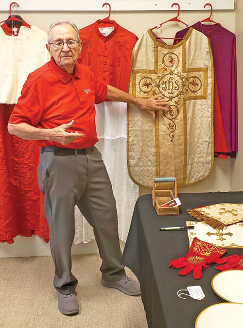 Ray Day, archivist for the Cardinal Ritter Birthplace Foundation, Inc., in New Albany points to a chasuble worn by Cardinal Joseph E. Ritter during his time as bishop (1934-1944) and archbishop (1944-1946) of Indianapolis. The long white vestment (mantelletta) hanging behind Day, the cape (rochet) hanging at far right, and the cream maniple and burse at the top of the table are also Ritter items from the same timeframe. The five pieces are on indefinite loan to the foundation from the Indianapolis Archdiocese&rsquo;s archives. The white ermine cape (mozzetta), scarlet cassock and scarlet mozzetta behind Day, as well as the liturgical gloves and china on the table, are a few of the Ritter items on indefinite loan from the St. Louis Archdiocese&rsquo;s archives.  (Photo by Natalie Hoefer)  